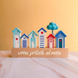 Può includere: Un ornamento decorativo in legno con sei piccole casette sulla spiaggia dipinte di blu, rosso e bianco. Ogni casetta ha dettagli unici come cuori e strisce. Una nuvola blu è sopra le casette e la base ha la scritta "vorrei portarti al mare."
