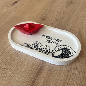 Könnte beinhalten: Weiße ovale Schale mit einem roten Papierboot und einem schwarz-weißen Wellendesign. Die Schale trägt den Text "In ogni mare insieme".