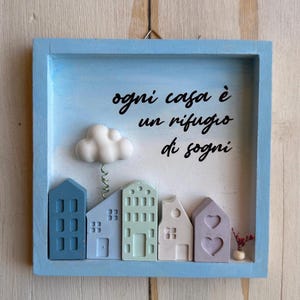Op de afbeelding: Een lichtblauw houten frame met een witte achtergrond en een citaat in het Italiaans dat luidt "ogni casa è un rifugio di sogni", wat vertaald wordt als "elk huis is een toevluchtsoord van dromen". Het frame bevat een 3D-scène van een klein stadje met huizen in verschillende pastelkleuren en een witte wolk met een veer.