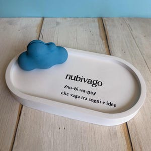 Può includere: Vassoio ovale bianco con una scultura di nuvola blu in cima. Il vassoio ha la parola "nubivago" scritta su di esso, insieme alla grafia fonetica e la frase "che vaga tra sogni e idee".