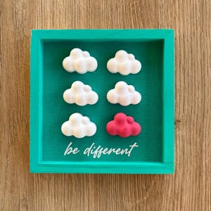 Op de afbeelding: Een turquoise omlijst kunstwerk toont zes wolkvormige objecten. Vijf zijn wit en één is felrood. De zin "be different" staat in witte letters onderaan. Het kunstwerk staat tegen een turquoise achtergrond.