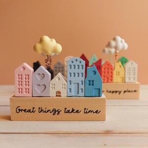 Può includere: Un paesaggio urbano in miniatura con case colorate e nuvole, su una base di legno con il testo "Great things take time".