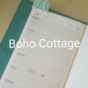 Puede incluir: Un planificador semanal con una pinza para encuadernar de color turquesa. El planificador tiene un fondo blanco con los días de la semana. Flechas decorativas están impresas en el planificador. El texto "Boho Cottage" está impreso.