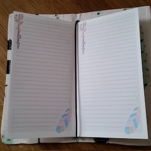 Może przedstawiać: Mały, biały notes z czarną oprawą i wzorzystą okładką. Okładka przedstawia wzór piór i strzał w odcieniach różowego, niebieskiego i zielonego.
