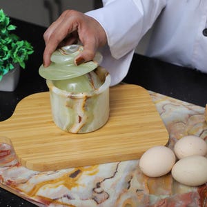 May include: A green and white onyx jar with a lid, on a wooden cutting board. The jar is cylindrical with a lid. The onyx has natural veining in green, white, and brown. The jar is being opened by a person wearing a white shirt.