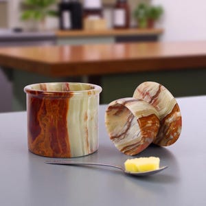 May include: A natural stone butter dish with a lid, showing a cylindrical base and a rounded lid. The dish has brown, cream, and green tones. A spoon holds a pat of butter.