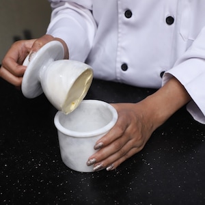 Könnte beinhalten: Ein weißer Butterbehälter aus Marmor mit Deckel, der Butter enthält. Die Schale ist rund mit einem zylindrischen Boden und einem Deckel mit einem flachen, kreisförmigen Griff. Der Butterbehälter steht auf einer schwarzen Oberfläche.