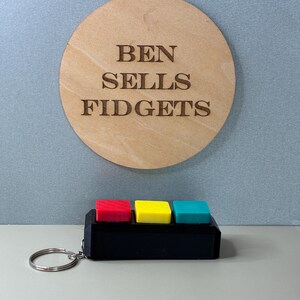 May include: A black fidget toy with three colored buttons: red, yellow, and teal. The toy has a silver keychain ring attached. Above the toy is a wooden circle with the text "BEN SELLS FIDGETS" engraved on it.