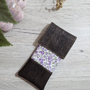 Può includere: Un astuccio rettangolare realizzato in sughero marrone scuro con una superficie testurizzata e un pannello centrale in tessuto bianco decorato con un piccolo motivo floreale viola. L'astuccio presenta una patta ripiegabile e cuciture visibili.