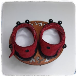 May include: A pair of red suede doll shoes with black trim and black beads. The shoes are on a wooden base with a decorative design.