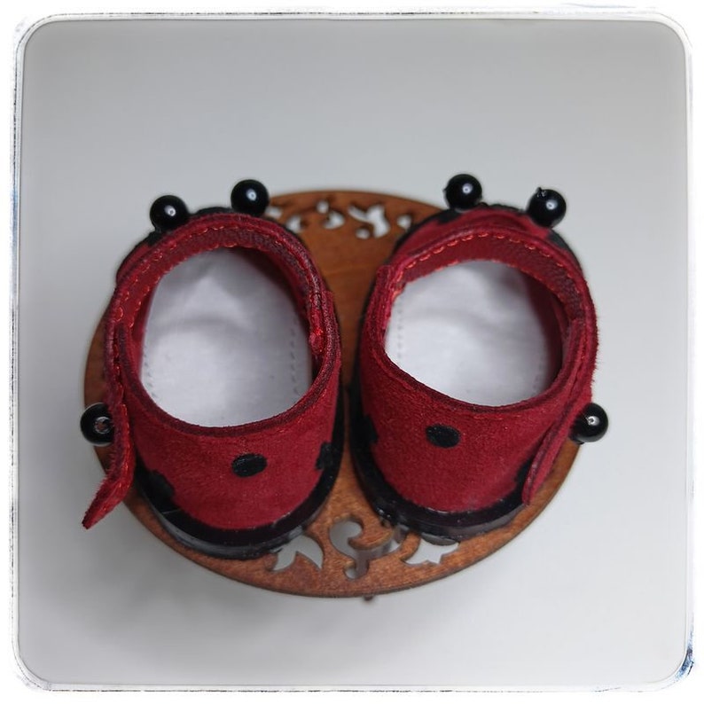 May include: A pair of red suede doll shoes with black trim and black beads. The shoes are on a wooden base with a decorative design.