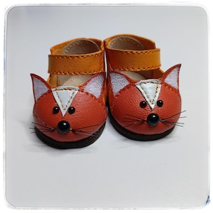 May include: A pair of orange and brown baby shoes with fox faces. The shoes have a strap closure and black whiskers. The fox faces have white accents and black button eyes. The shoes are made of leather.