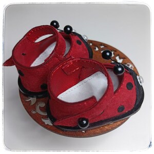 May include: A pair of red ladybug-themed shoes with black polka dots and black beads. The shoes are made of suede and have a black trim.
