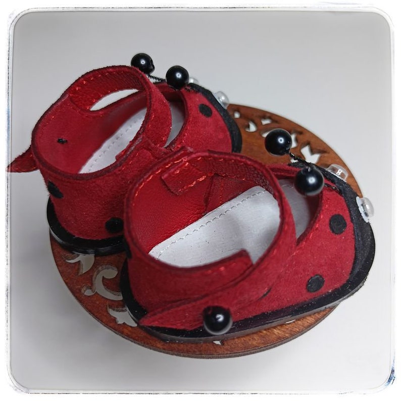 May include: A pair of red ladybug-themed shoes with black polka dots and black beads. The shoes are made of suede and have a black trim.