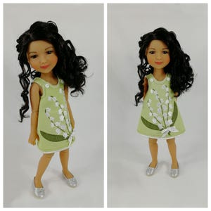 May include: Two dolls with dark curly hair and fair skin are shown. They wear light green dresses with white floral designs and white bows. The dolls have silver shoes and are posed against a white background.