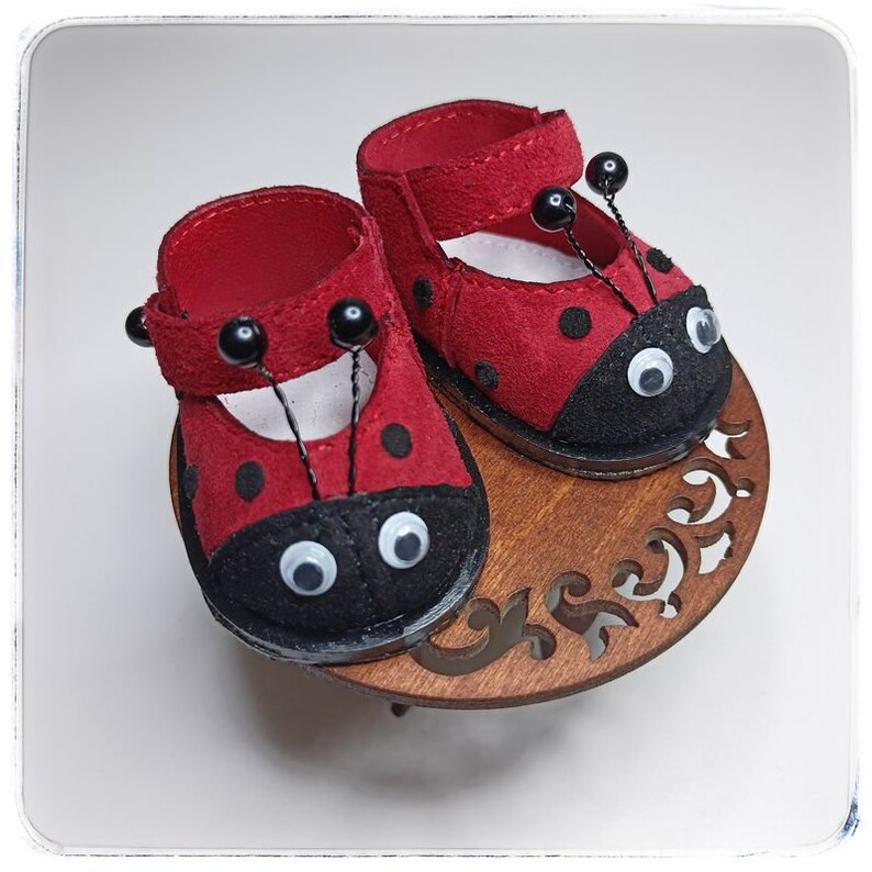 May include: A pair of red ladybug-themed shoes with black spots and white googly eyes. The shoes are on a wooden stand with a decorative cutout.