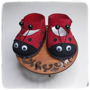 May include: A pair of red ladybug-themed baby shoes with black spots and white eyes. The shoes have black straps and are sitting on a wooden stand.