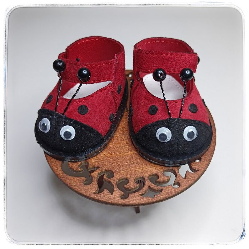 May include: A pair of red ladybug-themed baby shoes with black spots and white eyes. The shoes have black straps and are sitting on a wooden stand.