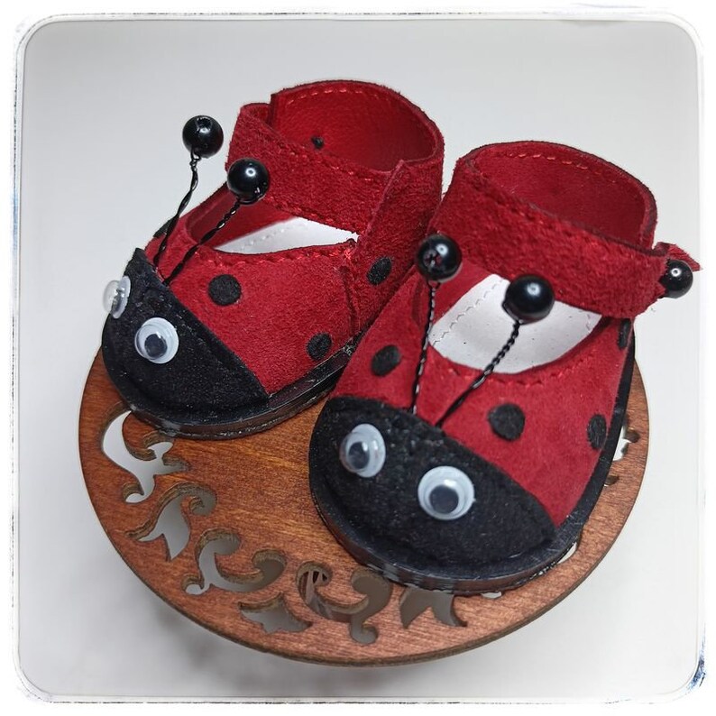 May include: A pair of red ladybug-themed shoes with black spots and white eyes. The shoes have black straps and are made of felt.