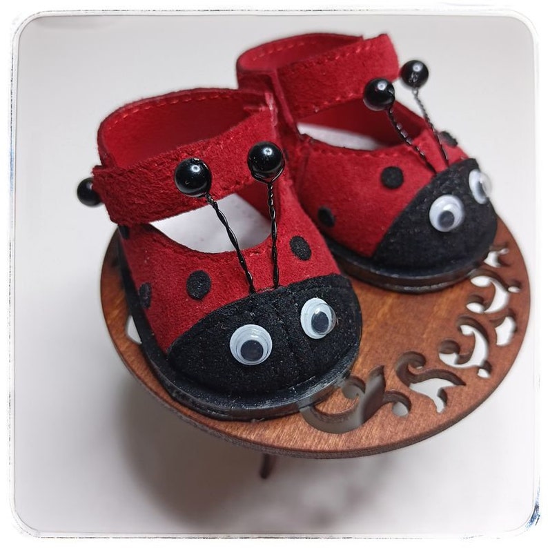 May include: A pair of red ladybug-themed shoes with black spots and white eyes. The shoes have black straps and are sitting on a wooden stand.