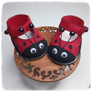 May include: A pair of red ladybug-themed doll shoes with black spots and white googly eyes. The shoes have black straps with black beads.