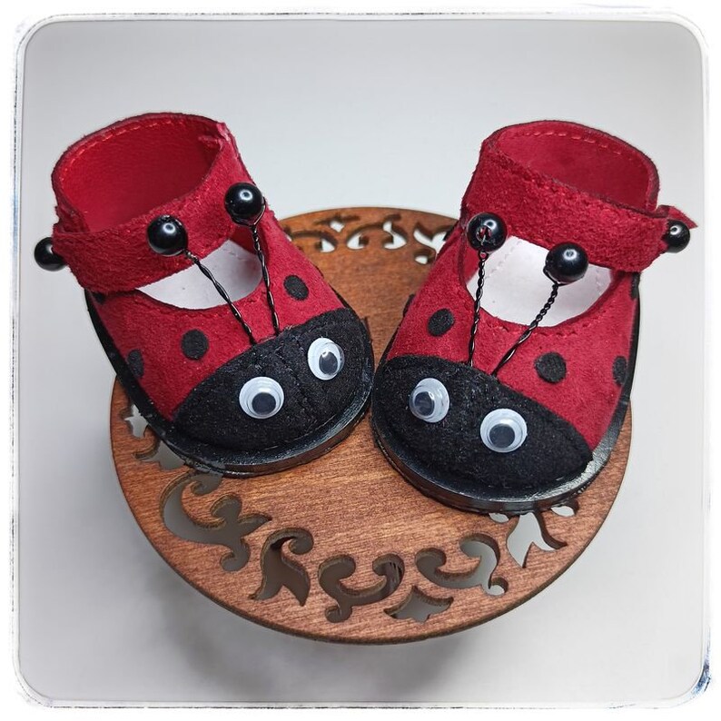 May include: A pair of red ladybug-themed doll shoes with black spots and white googly eyes. The shoes have black straps with black beads.