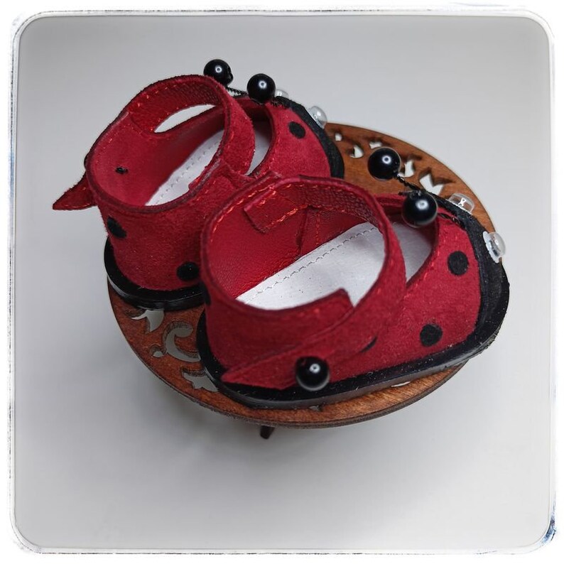 May include: A pair of red ladybug-themed shoes with black polka dots and black bead accents. The shoes are made of felt and have a black trim around the edges.