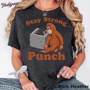 Könnte beinhalten: Dunkelgraues Melange-T-Shirt mit einem Cartoon-Orang-Utan und einem Baby-Affen mit den Worten "Stay Strong Punch". Der Orang-Utan ist braun und der Baby-Affe grau. Der Text ist in einem gebrannten Orange gehalten.