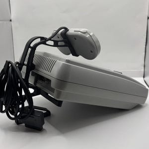 May include: A light grey video game console with a matching controller suspended above it by a black plastic mounting bracket. The controller is connected by a coiled black cord that hangs down from the side of the console unit.