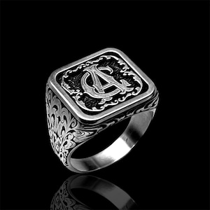 Può includere: Un anello sigillo color argento con una faccia rettangolare che presenta la lettera A incisa al centro. La superficie dell'anello è decorata con intricati motivi a volute che si estendono lungo i lati della fascia, creando un aspetto strutturato che contrasta con i dettagli scuri e incavati dello sfondo.
