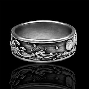 Puede incluir: Un anillo de plata con un diseño detallado de olas y una luna. El anillo es ancho y tiene una superficie texturizada.