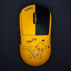 May include: A yellow and black computer mouse featuring a Pikachu design. The mouse has the word "Pokemon" printed on the top and the word "Yeah" with a heart and a Pikachu graphic on the bottom. The mouse is on a black surface.