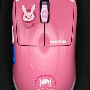 May include: Pink gaming mouse with white detailing and the text "I PLAY TO WIN". Features a bunny logo and the word "MEKA". Includes a scroll wheel and blue accents. A gaming accessory.