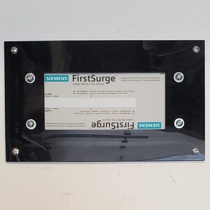 May include: A black rectangular panel with a white label that reads "FirstSurge" and "Surge Protective Device" with the Siemens logo. The panel is mounted on a white wall with silver screws.