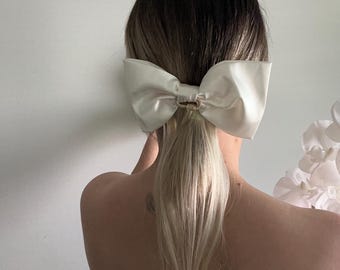 May include: A large, ivory satin bow hair accessory. The bow is secured with a gold-colored clasp and is worn at the base of a blonde ponytail. The background is a plain white wall.