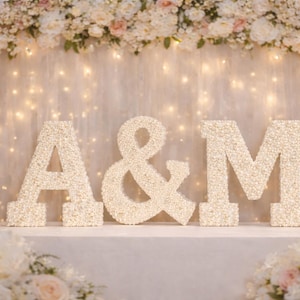 May include: A decorative floral display featuring the letters "A & M". The letters are made of white flowers and set against a backdrop of fairy lights and more floral arrangements. The scene evokes a celebratory atmosphere.