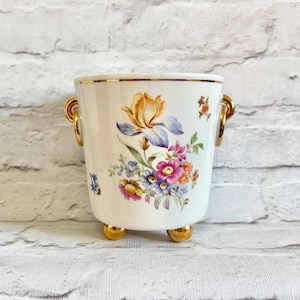 May include: A white ceramic planter with a gold rim, handles, and feet. The planter is decorated with a floral design featuring an iris and other colourful flowers. The planter is approximately 13 cm tall.