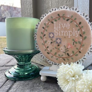 以下が含まれることがあります： 白い木製の刺繍枠に、ベージュの布地に「Live Simply」という文字が茶色の糸で刺繍されています。枠は白いスタンドの上に置かれ、手前に白い花が置かれています。