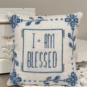 May include: Square, off-white pillow with blue cross-stitch embroidery. The pillow features the words "I AM BLESSED" in a blue frame. Floral and leaf patterns decorate the edges. The pillow is on a white crocheted surface.