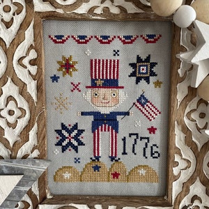 May include: A framed cross-stitch design featuring a cartoon Uncle Sam holding a flag and standing on clouds. The design includes stars, stripes, and the year 1776.