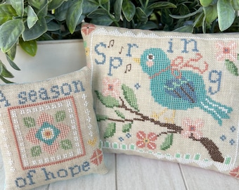 Könnte beinhalten: Zwei bestickte Kissenbezüge mit Frühlingsmotiv. Ein Kissen zeigt einen blauen Vogel auf einem Zweig mit rosa Blüten und dem Text "Frühling". Das andere Kissen zeigt ein rosa-blaues Blumendesign und den Text "Eine Zeit der Hoffnung".