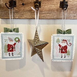 May include: Three handmade Christmas decorations featuring a Santa Claus design. The decorations are white with a blue border and a white pom-pom trim. The decorations are hanging from a wooden surface.