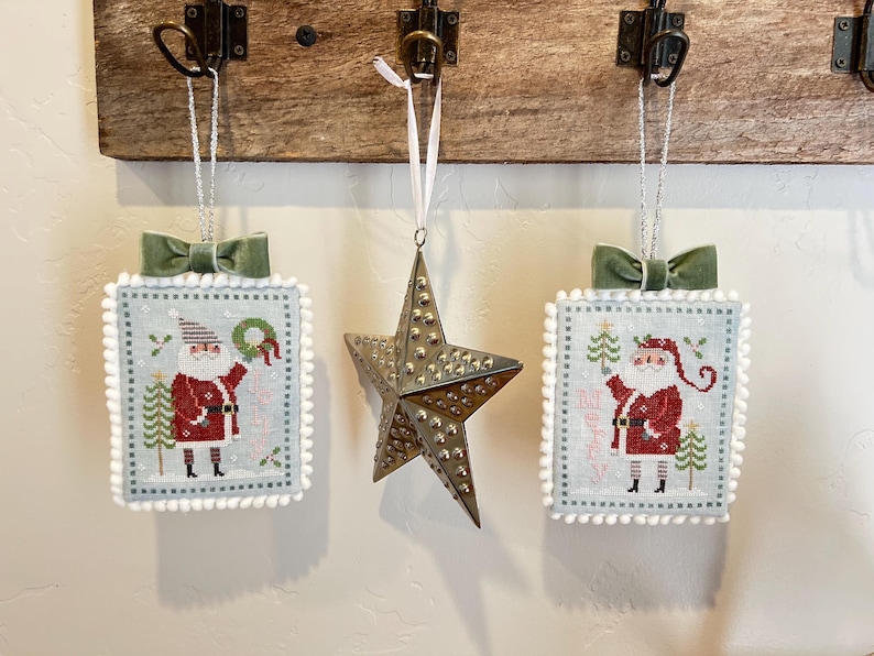 May include: Three handmade Christmas decorations featuring a Santa Claus design. The decorations are white with a blue border and a white pom-pom trim. The decorations are hanging from a wooden surface.