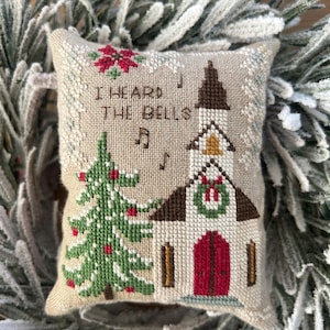 May include: A beige, square, cross-stitched Christmas ornament featuring a church, Christmas tree, and the text "I HEARD THE BELLS." The church has a red door and a green wreath. The ornament is surrounded by frosted greenery.