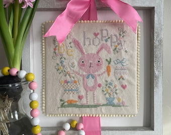 Könnte beinhalten: Ein gerahmtes Kreuzstich-Stickbild mit einem rosa Hasen und dem Text "Be Hoppy". Der Rahmen ist weiß und mit einem rosa Band verziert. Im Vordergrund befinden sich ein gelbes Holz-Küken und eine Perlenkette.
