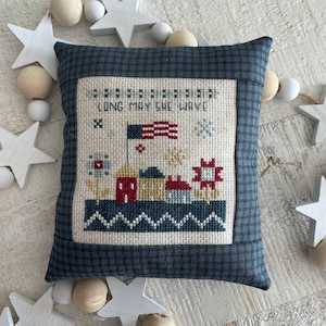 May include: A blue and white pillow with a cross-stitch design featuring a small town with houses, a flag, and the text "Long May She Wave".