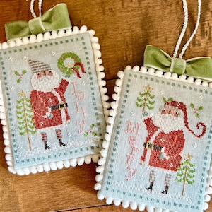 May include: Two blue and white cross-stitch ornaments with a white pom-pom trim. Each ornament features a Santa Claus figure with a red suit, white beard, and a green tree. The ornaments are decorated with green velvet ribbon bows and have the words "Merry" and "Jolly" stitched on them.