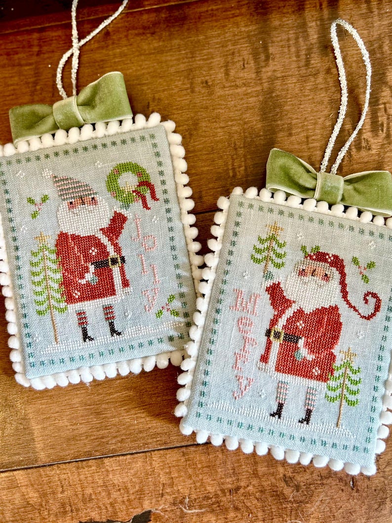 May include: Two blue and white cross-stitch ornaments with a white pom-pom trim. Each ornament features a Santa Claus figure with a red suit, white beard, and a green tree. The ornaments are decorated with green velvet ribbon bows and have the words "Merry" and "Jolly" stitched on them.