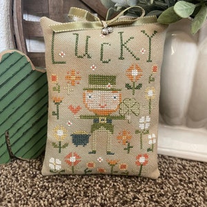 Può includere: Un cuscino in tessuto beige con un ricamo a punto croce che raffigura un folletto che tiene un pentolone d'oro. Il cuscino ha le parole "Lucky" ricamate in filo verde.