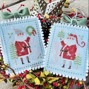 May include: Two blue and white cross-stitch ornaments featuring Santa Claus with the words "Jolly" and "Merry" on each ornament.